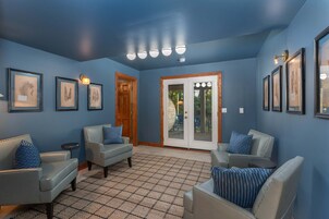A room with blue walls, a tiled floor, and framed botanical prints. There are two leather sofas with blue pillows, a wooden door, and a glass door leading outside.