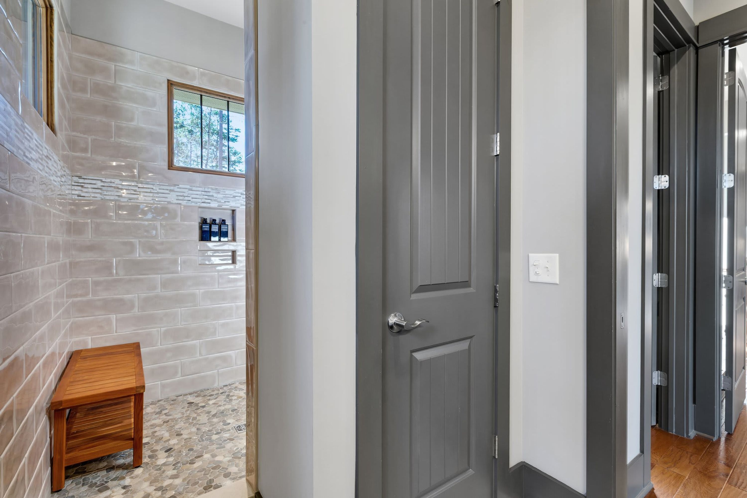 A hallway with a grey door, a small wooden bench, and a tiled wall with a window.