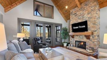 A spacious living room with a stone fireplace, a television mounted on the wall, and a large window overlooking a deck.