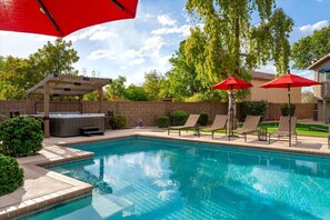 Una piscina con sillas de descanso y sombrillas, un jacuzzi y una pérgola.