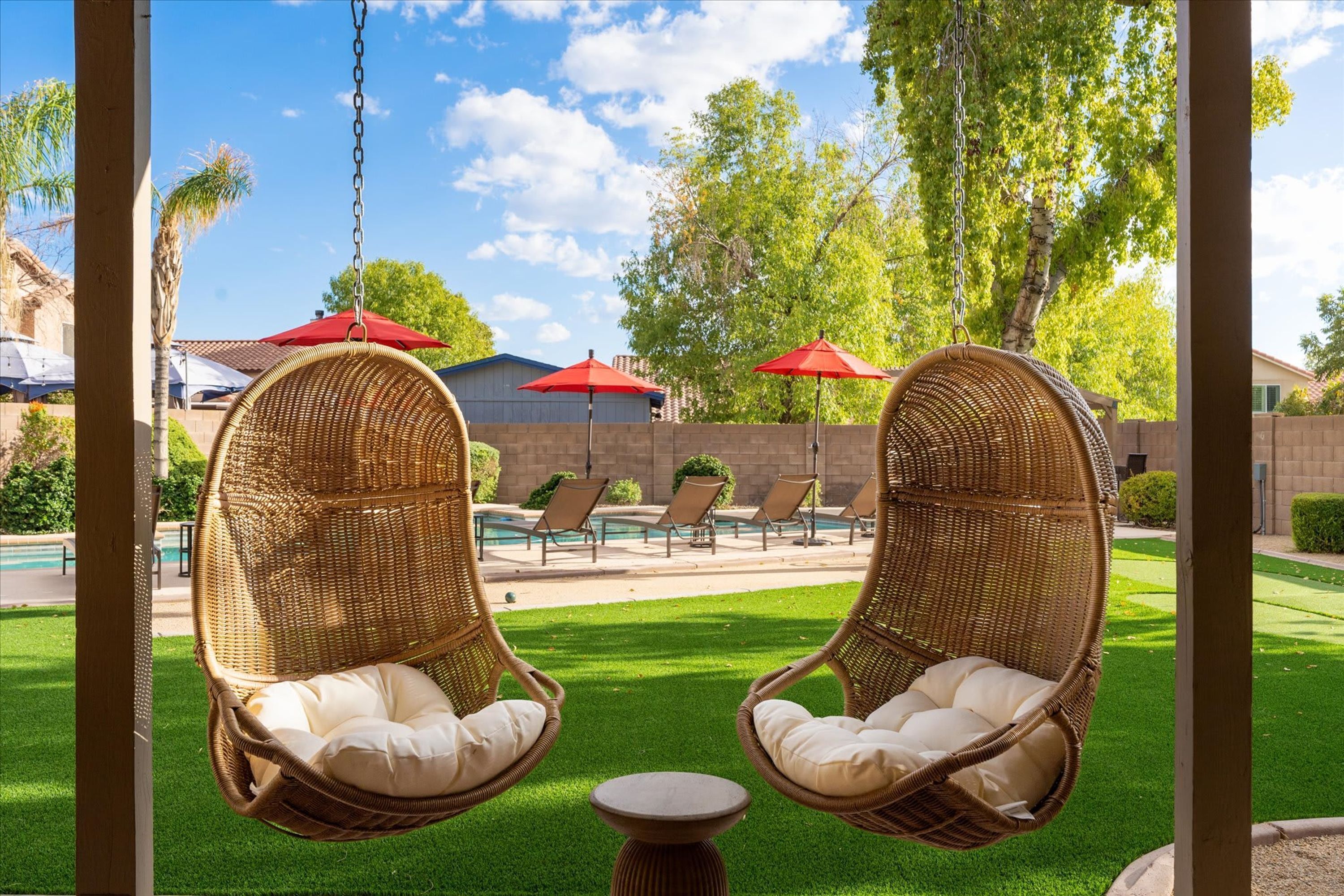 Deux fauteuils à bascule en osier avec des coussins, une piscine, des chaises longues et des parasols rouges, le tout dans un cadre extérieur ensoleillé.