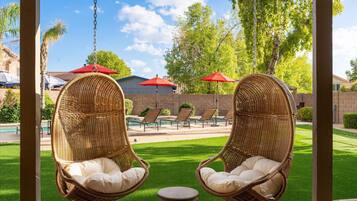 Deux fauteuils à bascule en osier avec des coussins, une piscine, des chaises longues et des parasols rouges, le tout dans un cadre extérieur ensoleillé.