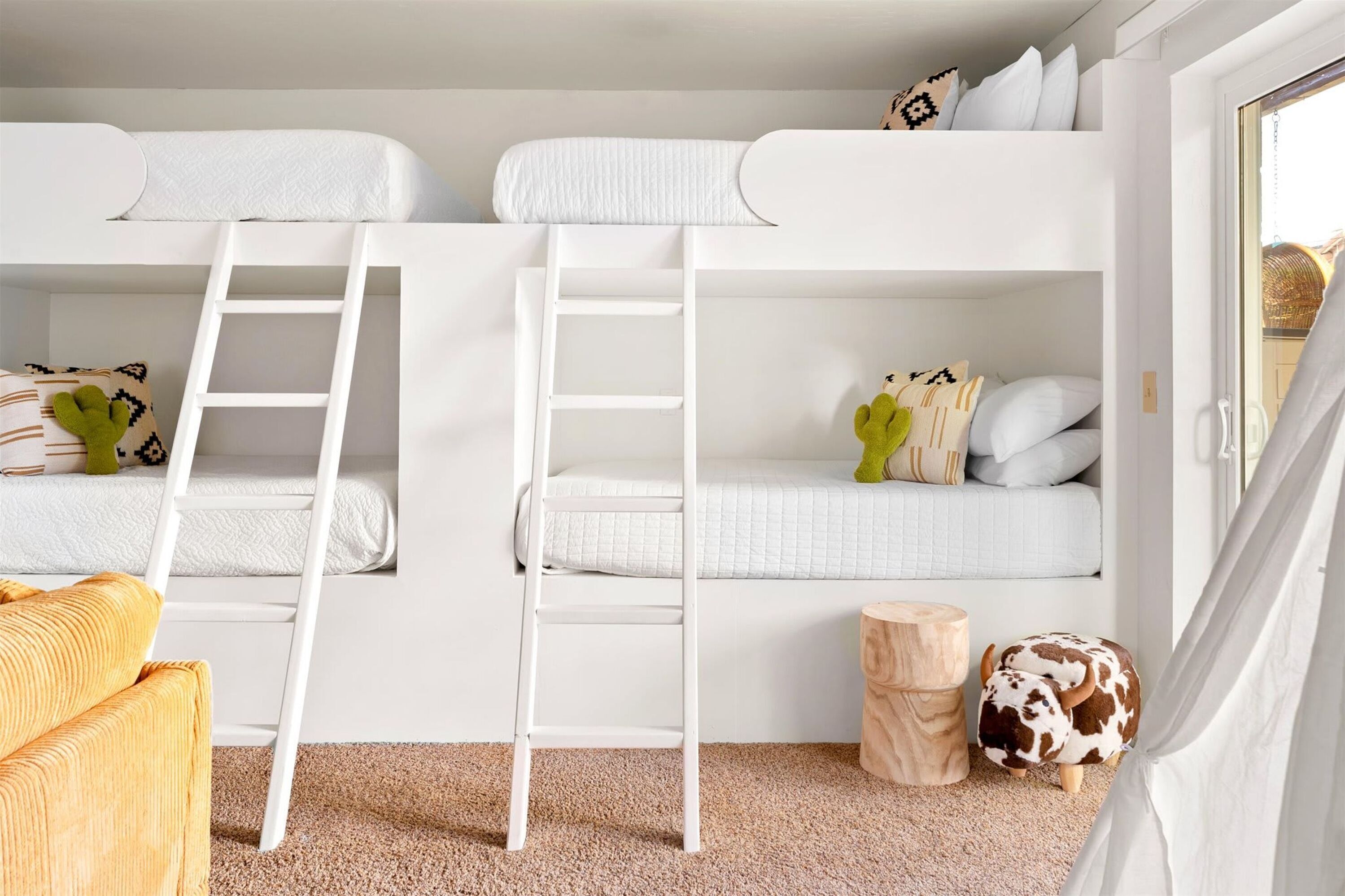 Habitación con literas, una escalera, un taburete de madera y un cojín estampado con vacas.
