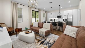 Un salon moderne avec un canapé, un pouf et un coin repas, la cuisine étant visible à l’arrière-plan.