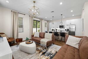 Ein modernes Wohnzimmer mit Sofa, Hocker und Essbereich, im Hintergrund eine Küche.