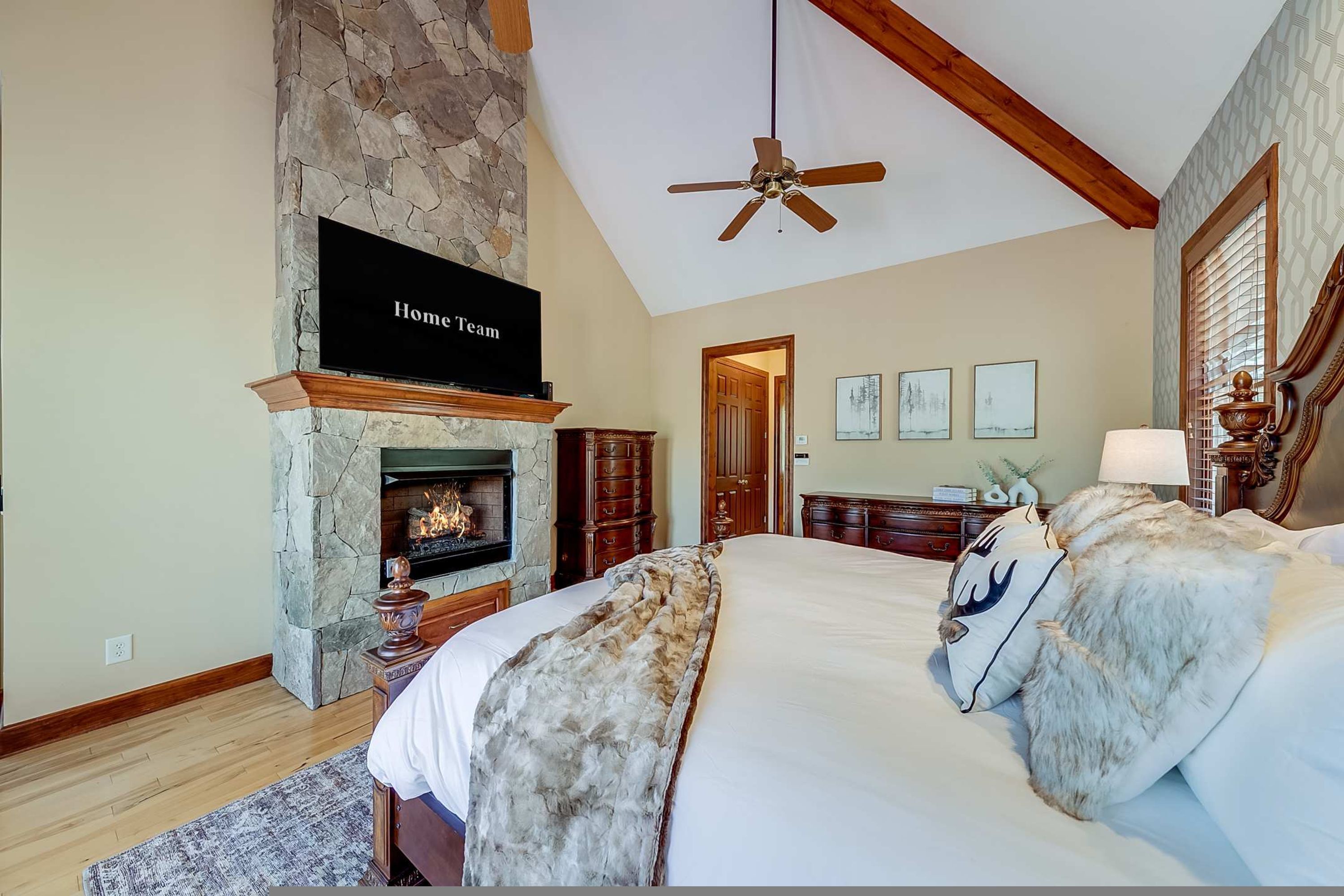 A bedroom with a stone fireplace, a ceiling fan, and a bed with a fur throw.