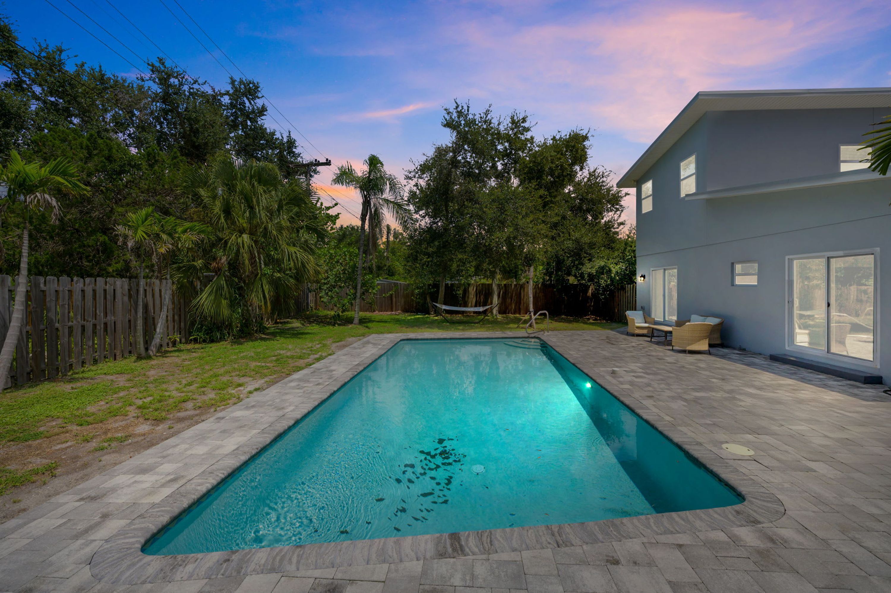 A rectangular swimming pool with a stone border, surrounded by a wooden fence, a grassy lawn, and a house with large windows.