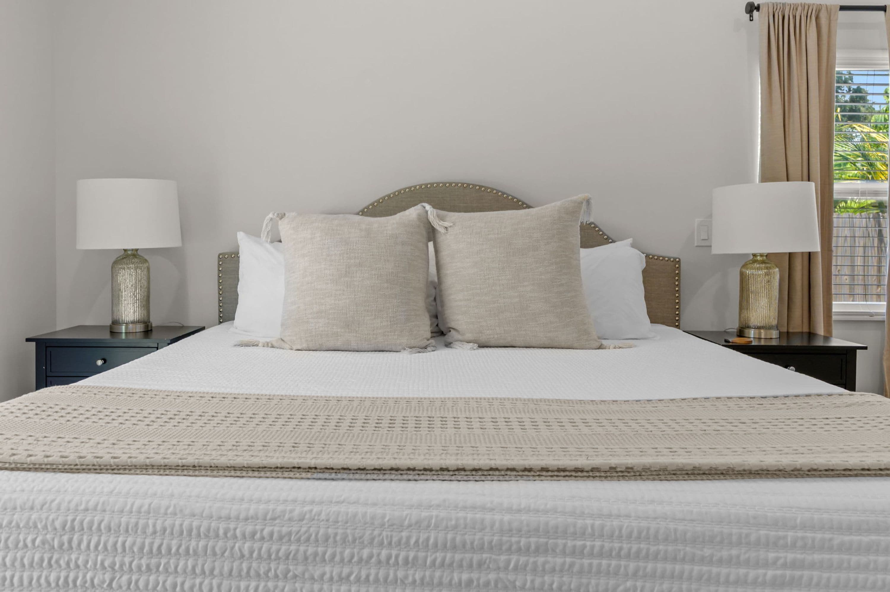 A neatly made bed with a headboard, two bedside tables with lamps, and a view of greenery outside.