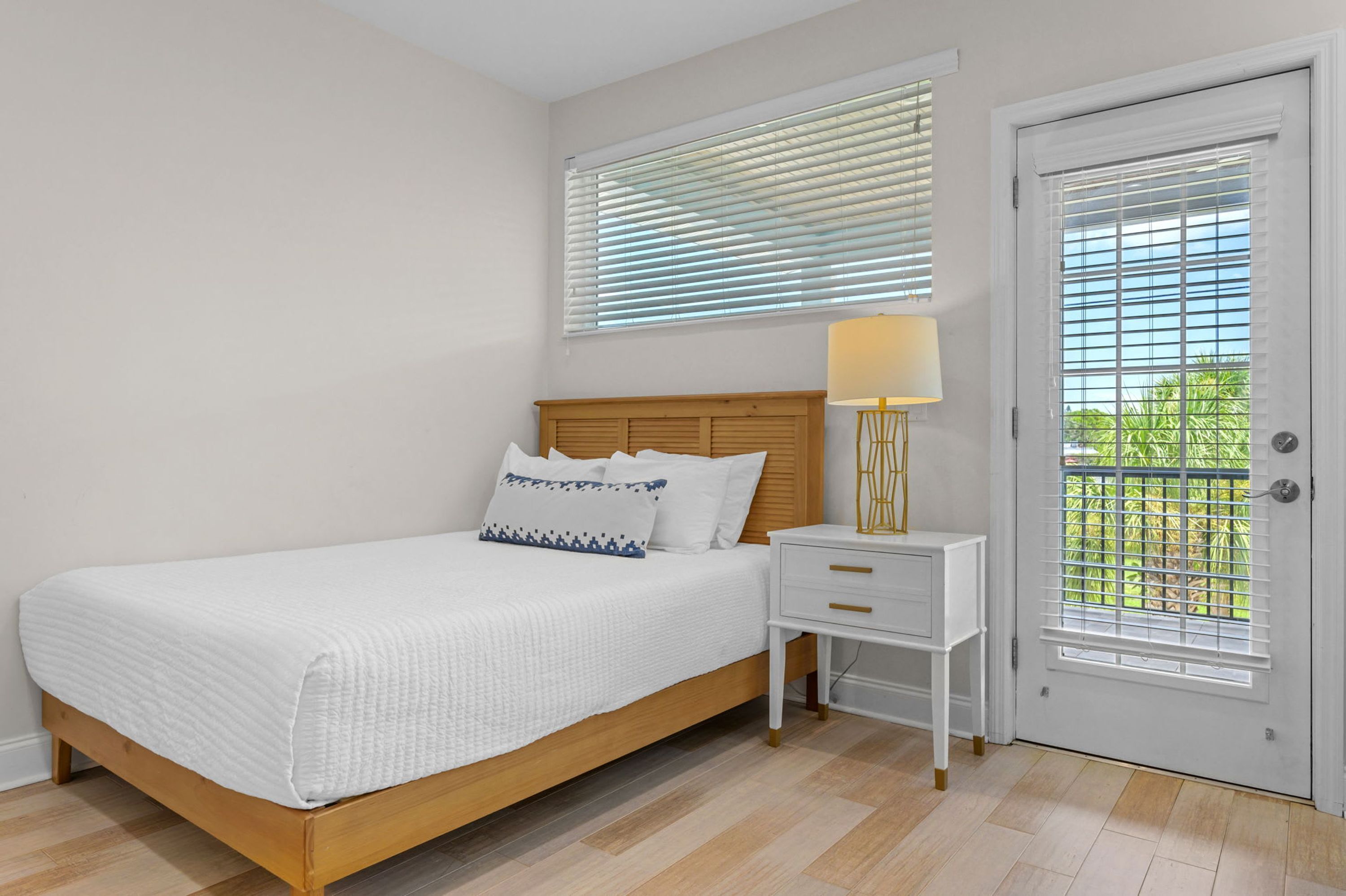 A bedroom with a bed, bedside table, lamp, and a door with blinds.