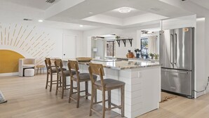 Una cocina moderna con una isla central, taburetes de bar, un refrigerador grande y un comedor con una silla y una mesa pequeña.