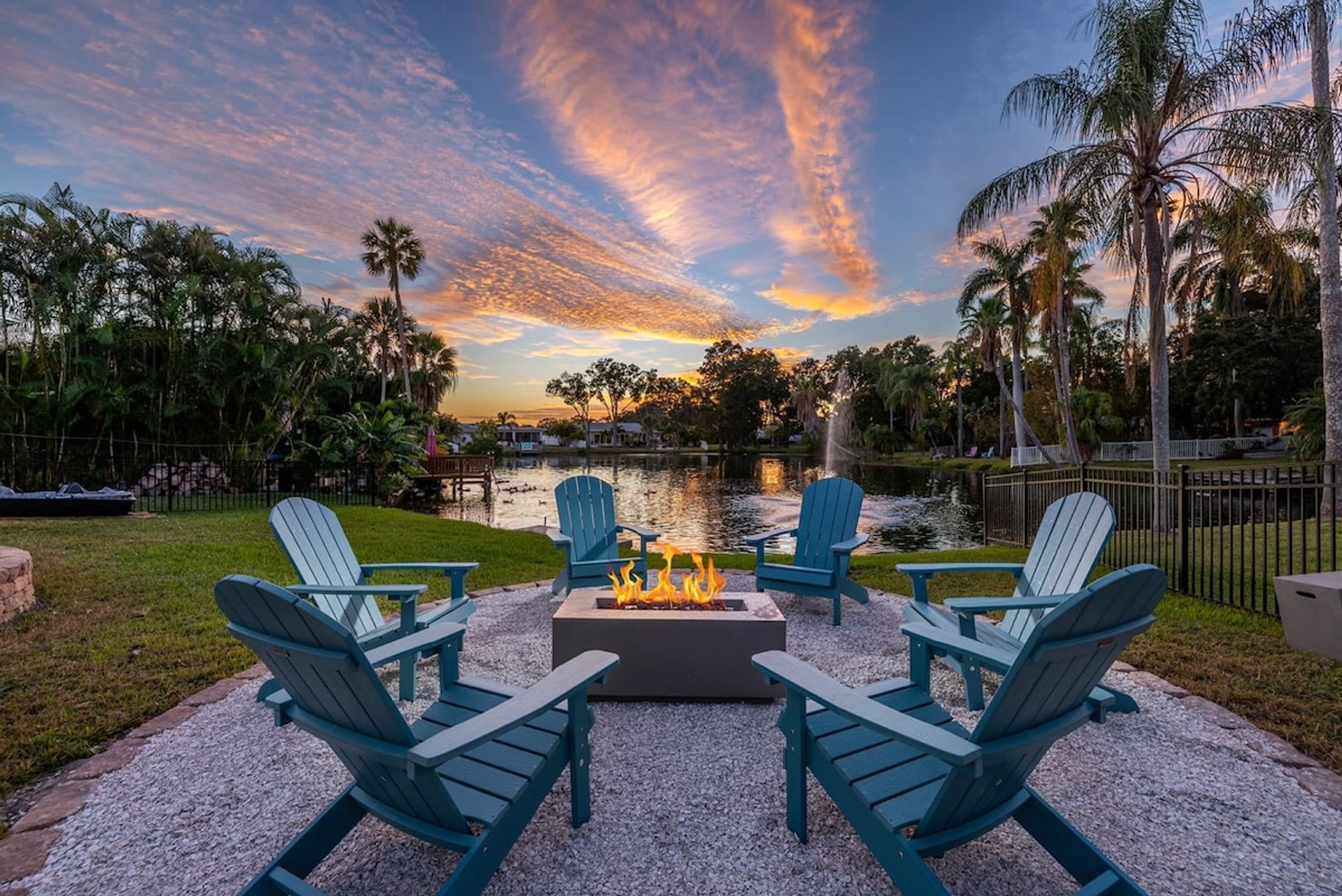 Un espace extérieur aménagé avec des chaises Adirondack et un foyer, offrant une vue sur un plan d'eau bordé de palmiers et illuminé par un ciel au coucher du soleil.