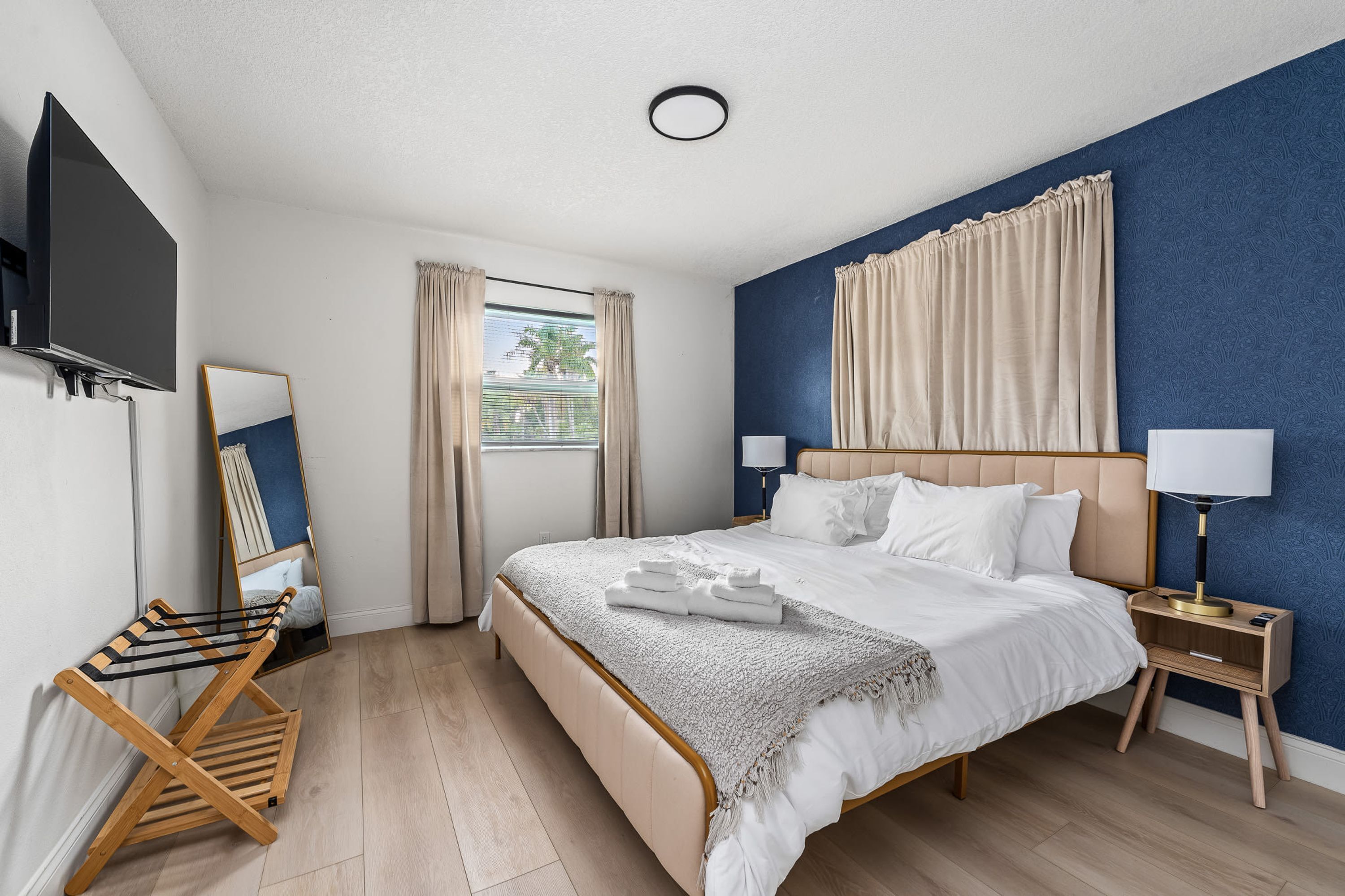 A hotel room with a bed, bedside tables, a TV, a window with curtains, and a mirror.