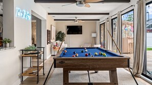 Una mesa de billar con bolas, un ventilador de techo, un televisor y una ventana con cortinas.