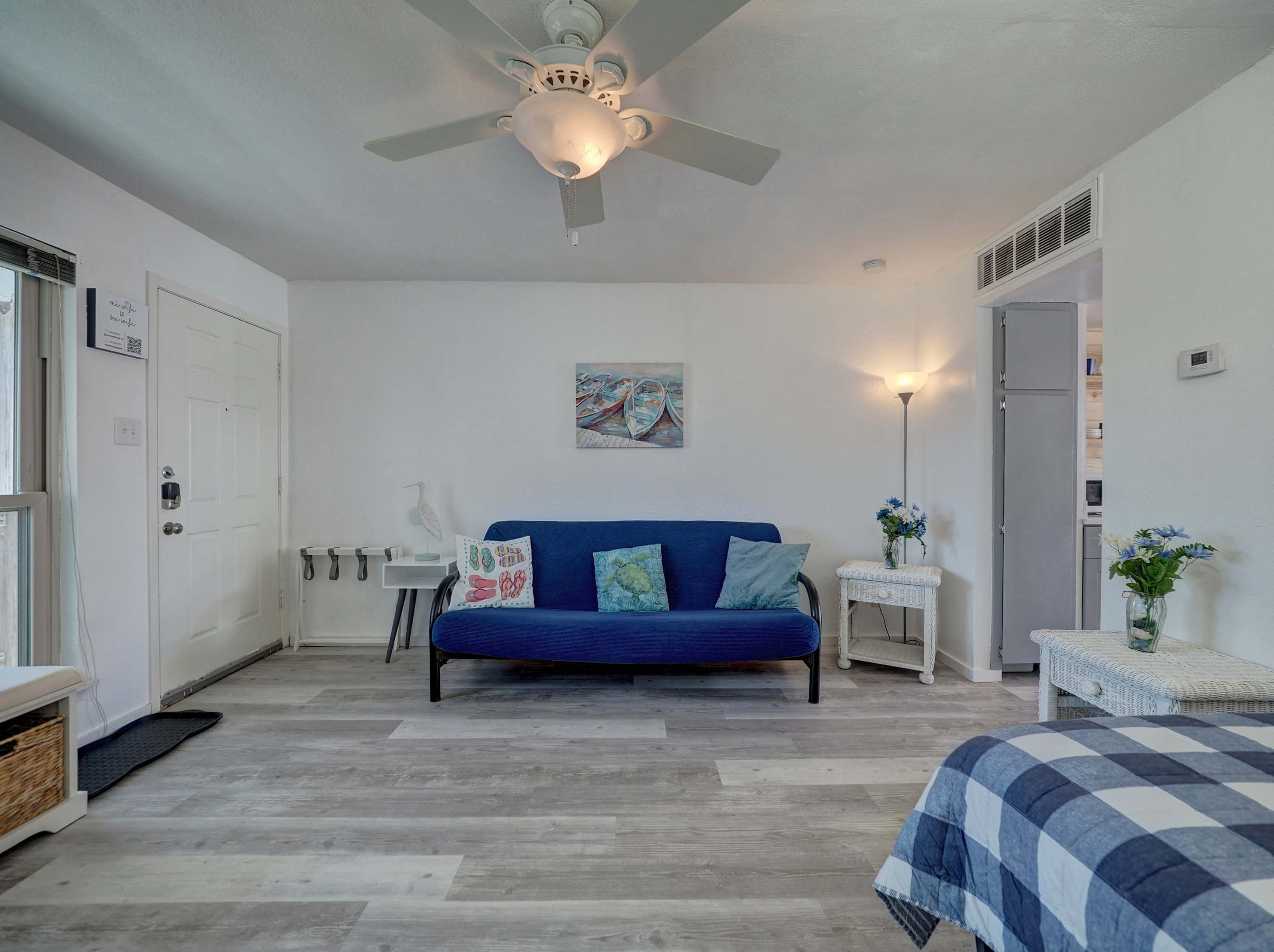 Ein Schlafzimmer mit einem blauen Sofa, einem Deckenventilator, einem Gemälde, einem Nachttisch mit Lampe und einem Bett mit kariertem Überwurf.