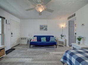 A bedroom with a blue sofa, a ceiling fan, a painting, a bedside table with a lamp, and a bed with a checkered blanket. - Waterfront Condo 1 Block to Beach Pool Access (Corpus Christi)