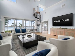 A spacious living room with a large TV, a staircase, and a view of the water. - Luxe Lakefront w Pool Pickleball Private Dock (Sebring)
