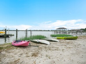 A sandy beach with kayaks and paddleboards, a dock with a covered structure, and calm water. - Luxe Lakefront w Pool Pickleball Private Dock (Sebring)