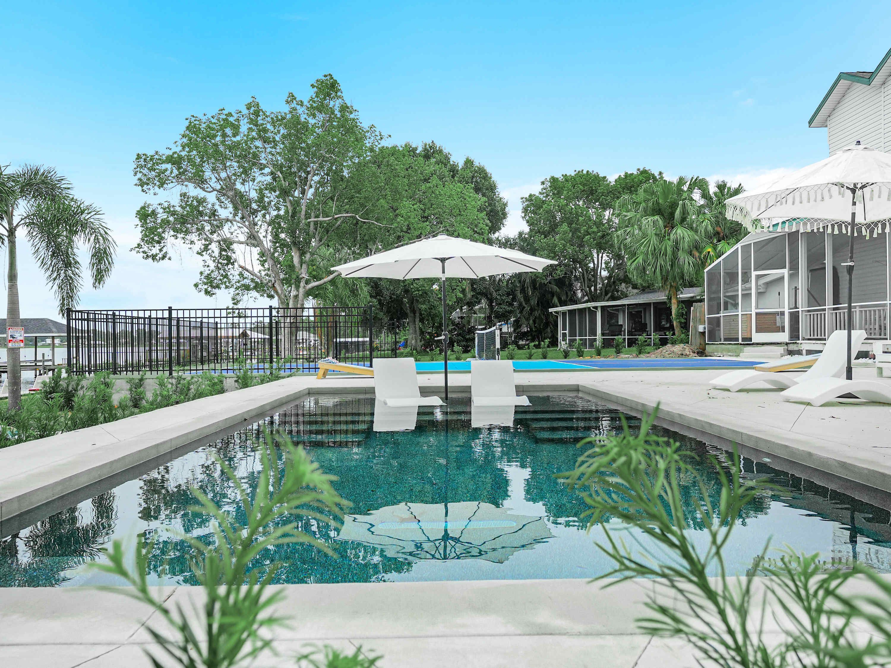 A swimming pool with lounge chairs and umbrellas, surrounded by greenery and a fence.