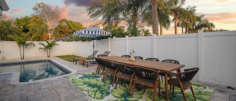 Un patio trasero con piscina, una mesa de madera con sillas y una valla blanca.