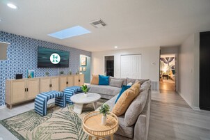 Uma sala de estar moderna com um sofá, pufes, uma mesa de centro e uma TV fixada numa parede estampada.