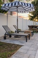 Un patio con dos sillones de descanso, una sombrilla a rayas y un reposapiés de mimbre.