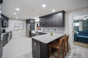 Una cocina moderna con armarios oscuros, una isla central y una zona de estar con un sofá azul y sillas de madera.