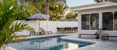 A swimming pool with lounge chairs and a table, surrounded by a white fence and trees.