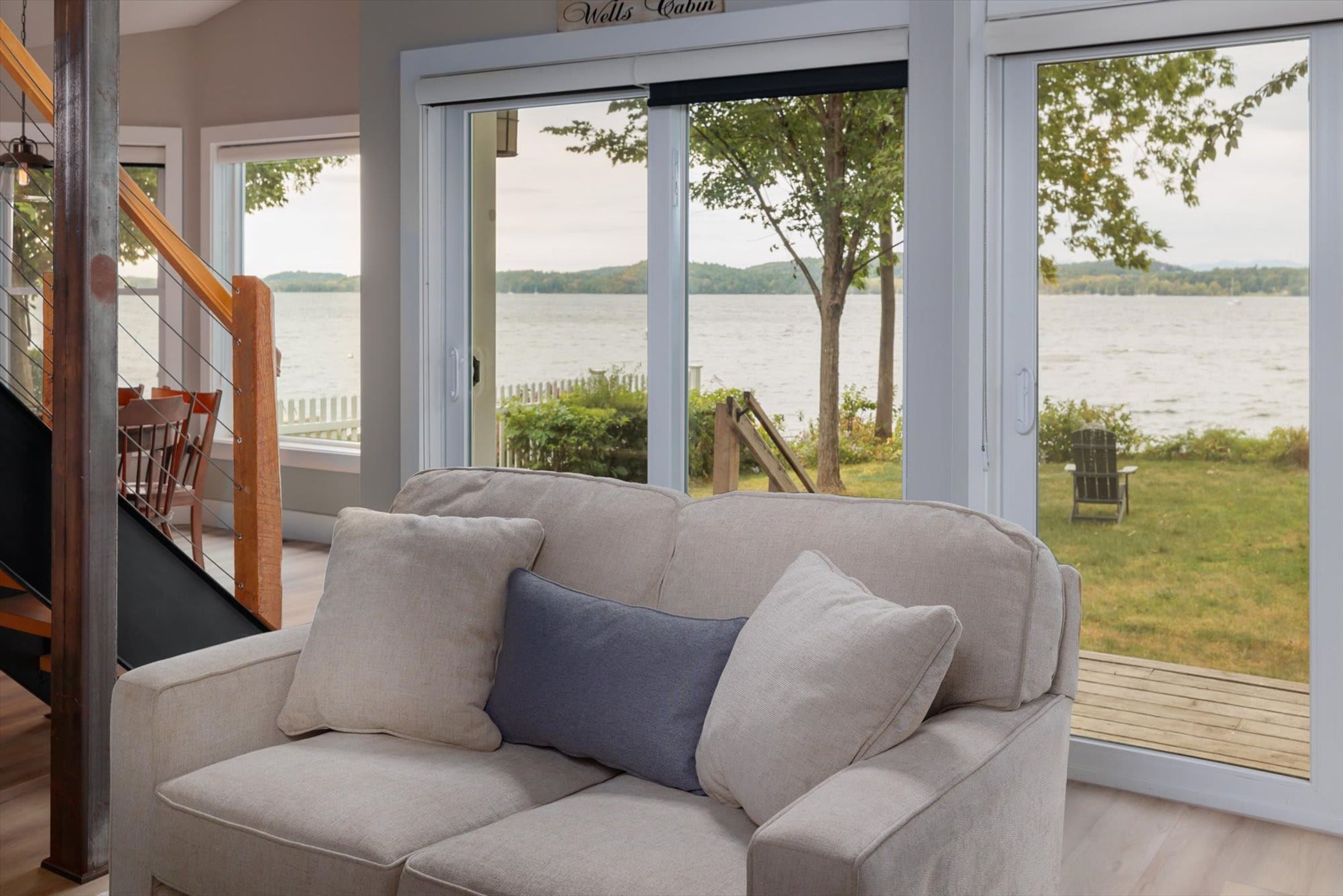 A room with a beige sofa, a wooden staircase, and a view of a lake through large glass doors.
