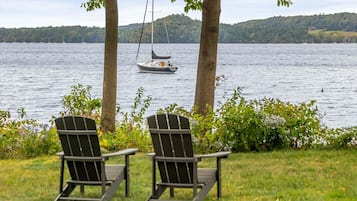 Two Adirondack chairs facing a lake with a sailboat in the distance and a forested hill in the background.