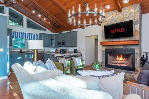 Uma sala de estar moderna com lareira, uma televisão montada exibindo 'NETFLIX' e uma cozinha ao fundo.