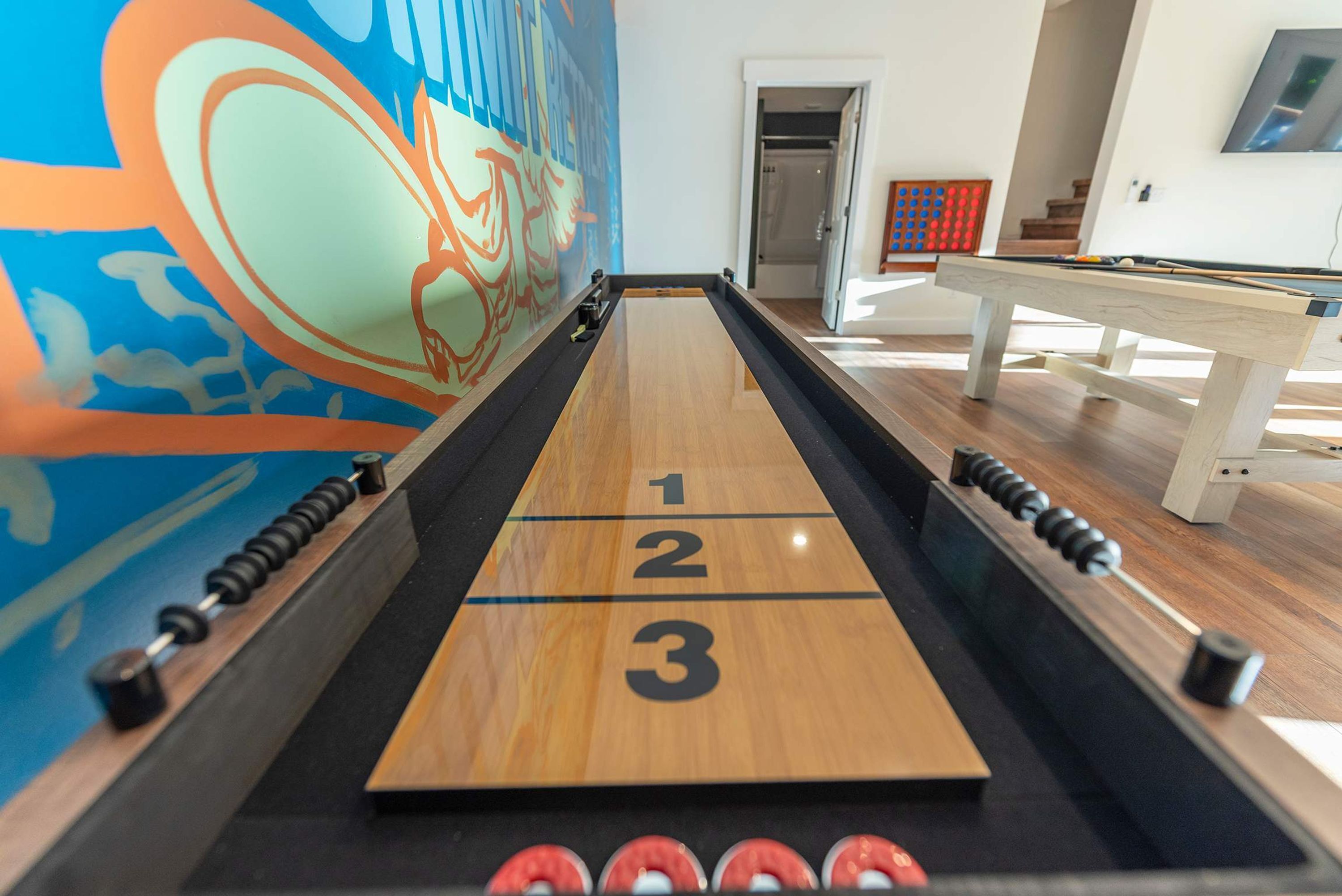 A shuffleboard table with numbered ends, a pool table, and a colorful mural in the background.