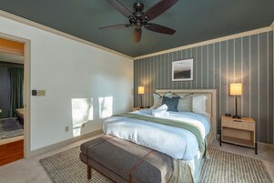 Quarto com cama, ventilador de teto, duas luminárias de cabeceira, mesa de cabeceira, um banco e um quadro na parede.