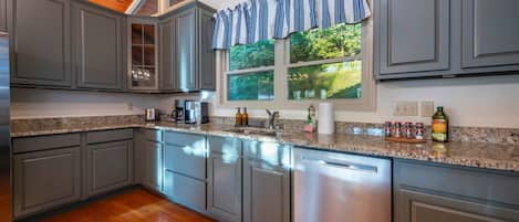 A kitchen with grey cabinets, a granite countertop, a stainless steel dishwasher, and a window with a view of greenery.