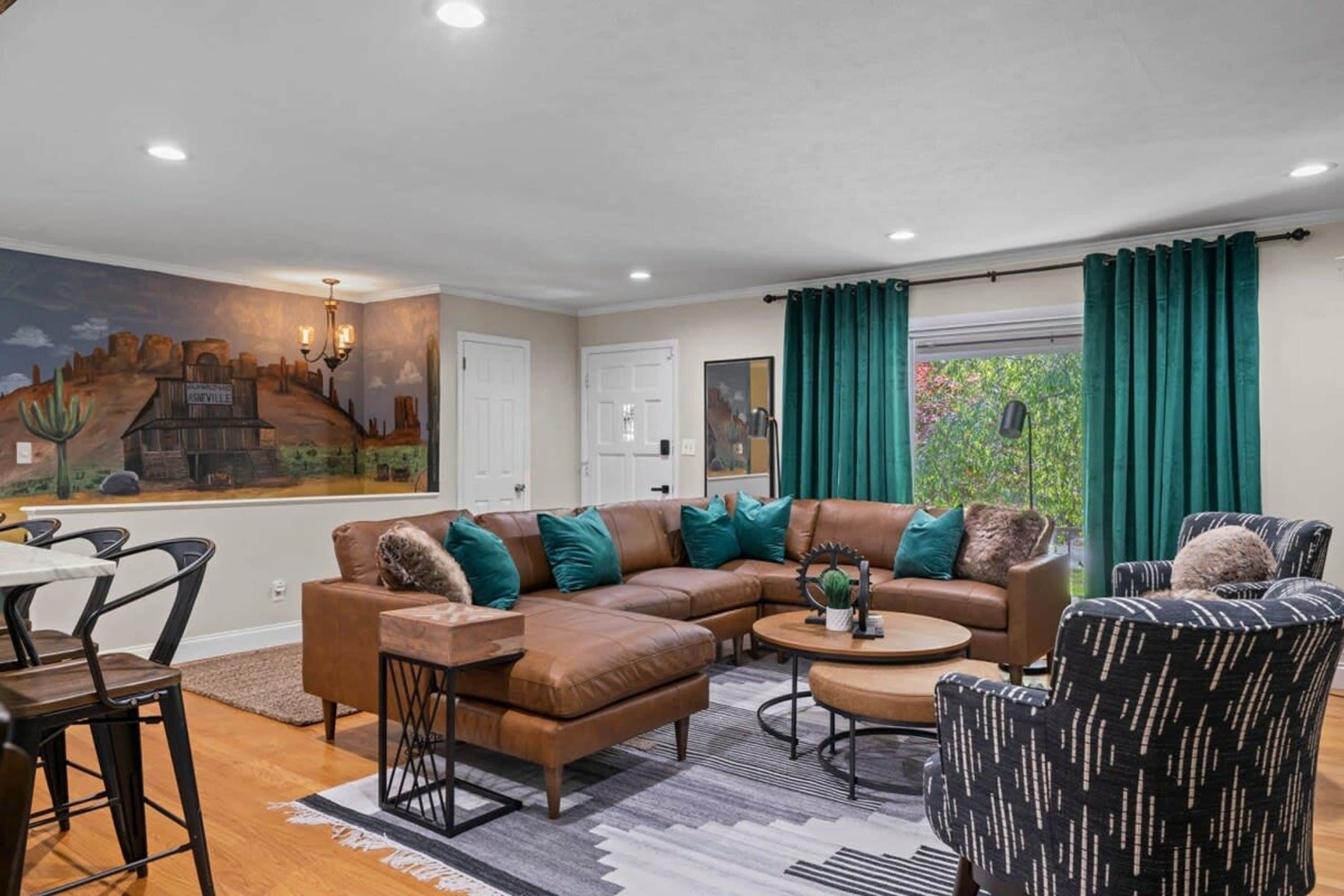 A living room with a sectional sofa, a coffee table, and a patterned armchair. There is a painting on the wall and a window with curtains.