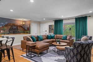 Ein Wohnzimmer mit einer Eckcouch, einem Couchtisch und einem gemusterten Sessel. An der Wand hängt ein Gemälde und es gibt ein Fenster mit Vorhängen.