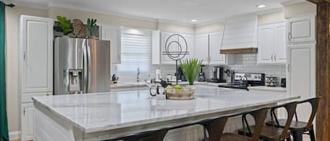 A modern kitchen with white cabinetry, a stainless steel refrigerator, a marble island with bar stools, and a built-in oven.