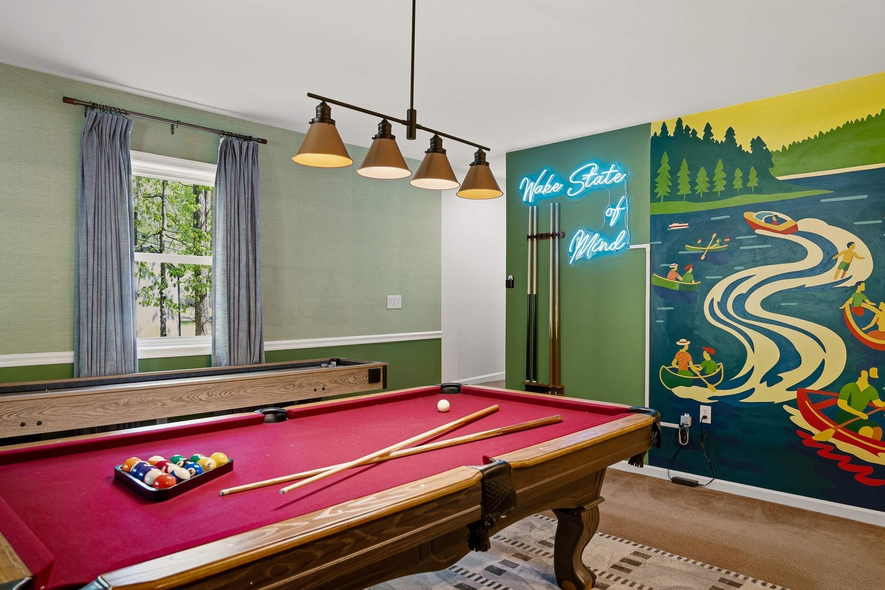 A pool table with cues and balls, a neon sign, and a mural of a river scene.
