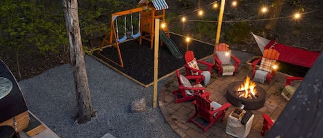 Un patio trasero con jacuzzi, un área de estar exterior con sillas Adirondack, un conjunto de columpios y una chimenea.