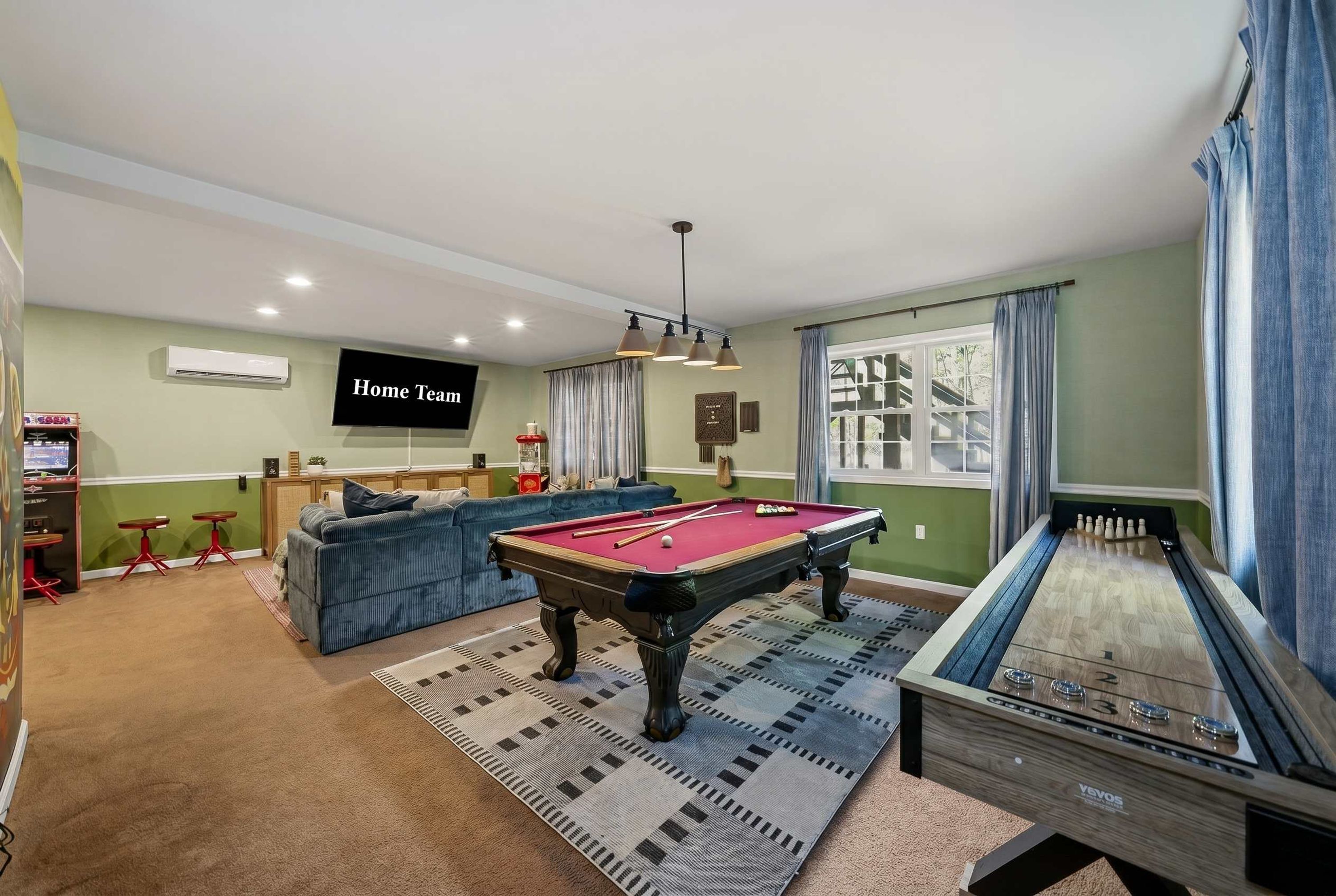 A room with a pool table, a snooker table, a sofa, a television, and a bar area with arcade games.