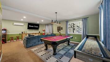 A room with a pool table, a snooker table, a sofa, a television, and a bar area with arcade games.