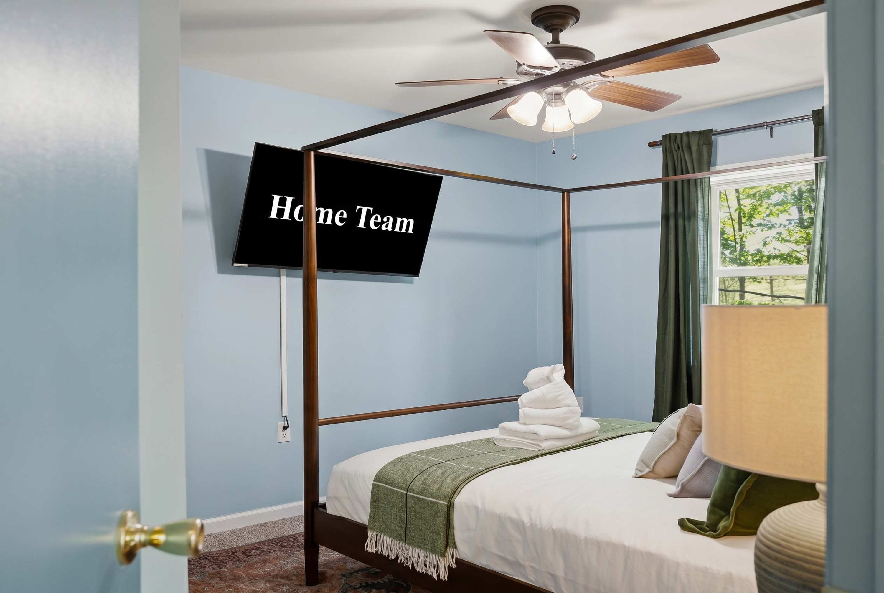 A four-poster bed with a television mounted on the wall, a ceiling fan, and a window with curtains.