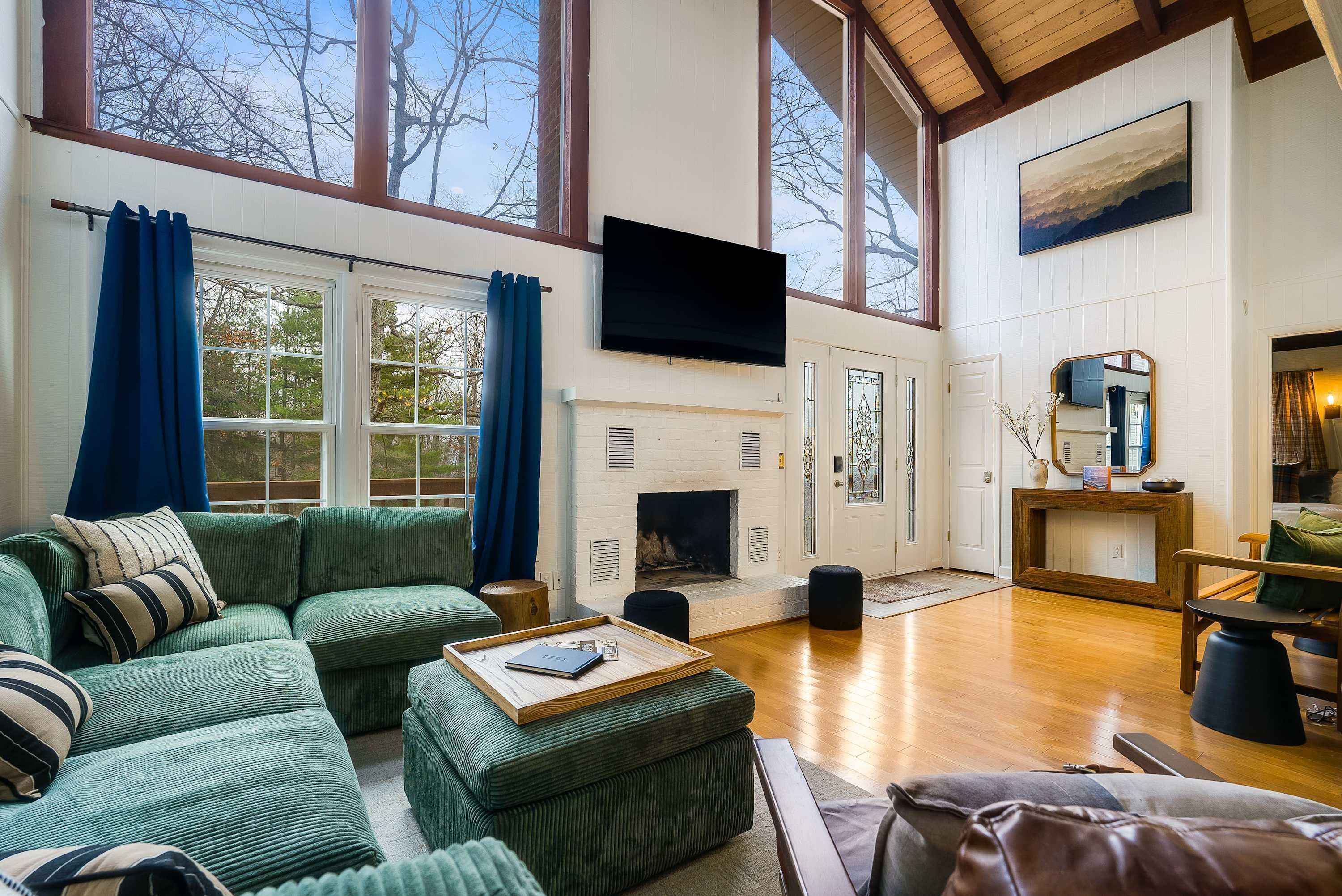 A spacious living room with a fireplace, a television mounted on the wall, and large windows with blue curtains.