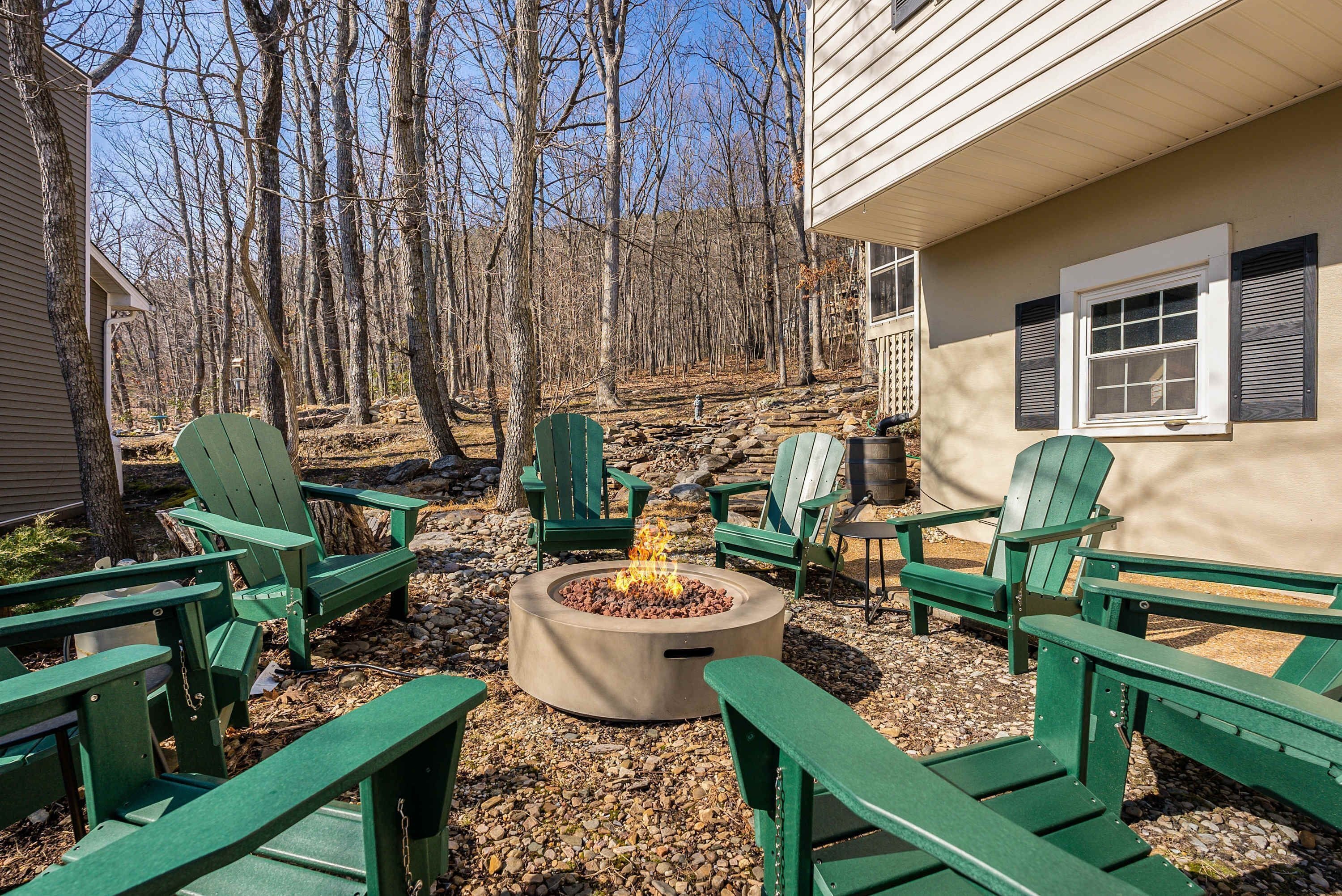 A backyard with a fire pit surrounded by Adirondack chairs, a stone wall, and a house with a window and shutters.