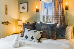 A hotel room with a bed, pillows, a nightstand with a lamp, a fish decoration, and a window with a view of trees. - Cozy Retreat - Sauna Hot Tub Pool Pass Game Room (McGaheysville)
