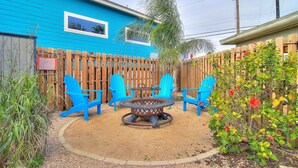 A backyard with a blue house, a wooden fence, and outdoor furniture including chairs and a table. - Sunshine Cottage- Fire Pit Fenced in Yard (Port Aransas)