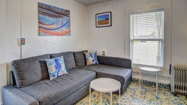 A living room with a grey sofa, two patterned throw pillows, a small white table, and a window with blinds. - Sunshine Cottage- Fire Pit Fenced in Yard (Port Aransas)