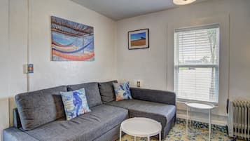 Una sala de estar con un sofá gris, dos cojines decorativos estampados, una mesita blanca y una ventana con persianas.