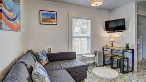 A living room with a grey sofa, a small round table, a TV mounted on the wall, and a window with blinds. - Sunshine Cottage- Fire Pit Fenced in Yard (Port Aransas)