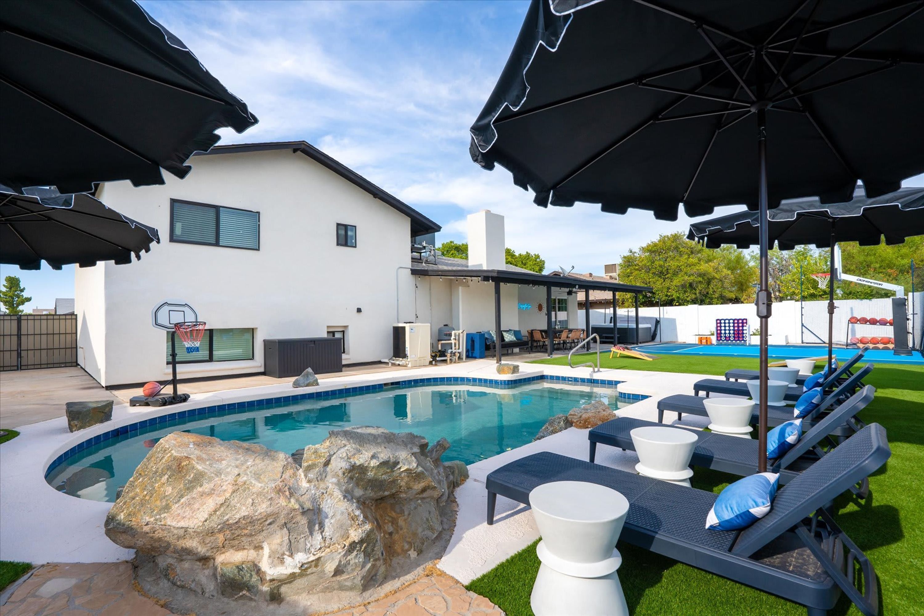A swimming pool with lounge chairs and umbrellas, a basketball hoop, and a house with a covered patio.