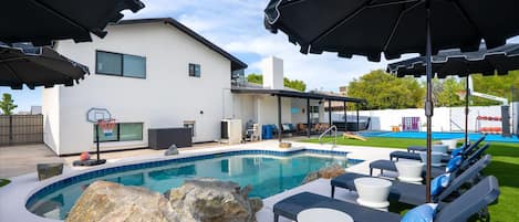 A swimming pool with lounge chairs and umbrellas, a basketball hoop, and a house with a covered patio.
